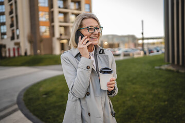 One mature woman outdoors in front of buildings talking on mobile phone and holding a to-go coffee, modern business and lifestyle concept