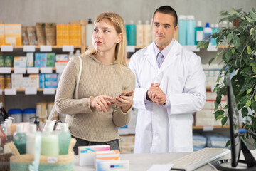 Obraz premium Woman scrolls through online catalog on the pharmacy website against the background of a pharmacist. Customer holds a mobile phone in hands in a pharmacy and looking at shelves with goods