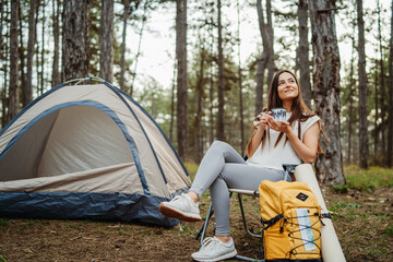 Young caucasian woman hiker enjoy hot drink near her tent in scenic forest	

