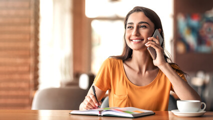 Cheerful young lady having conversation with employer on mobile phone while having coffee break at...