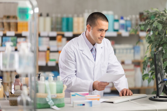 Man pharmacy employee works at computer, reads handbook on pharmaceutical care and additional literature, improves competence. Pharmacist during work, office equipment and modern technologies