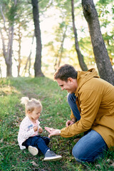 Dad holds out a tiny mushroom in the palm to a little girl sitting in the forest on the grass