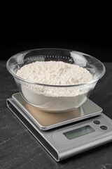 Electronic kitchen scale with bowl of flour on dark textured table against black background, closeup