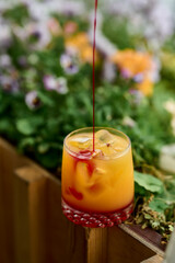 Cocktail in a glass with ice against a background of flowers and syrup pouring from the top