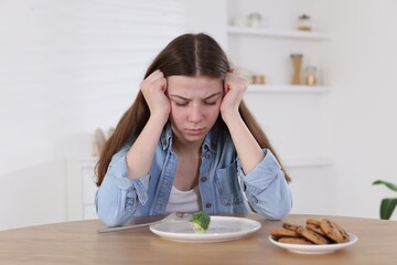 Sad teen girl at table with broccoli indoors. Eating disorder