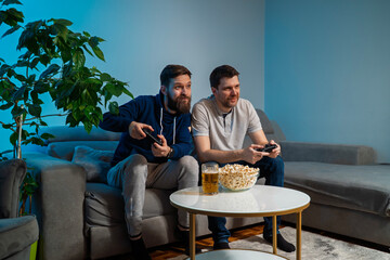 Two Caucasian guys playing video games at home drinking beer and eating popcorn 