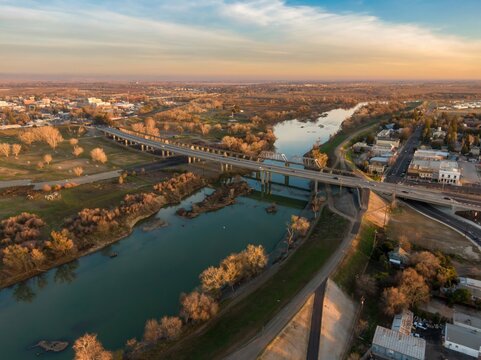 Aerial view of Yuba City, California, USA, at sunset. The Feather River flows under bridges carrying cars and pedestrians. The image shows the city's layout and transportation.