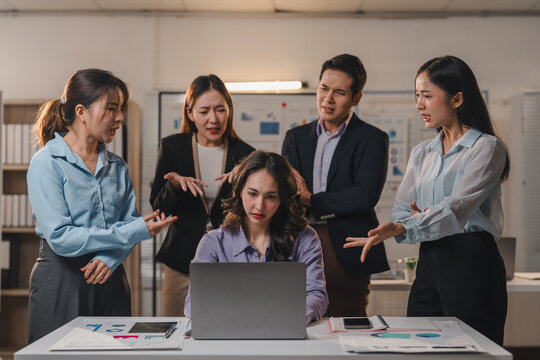 Stressed businesswoman working on a laptop in a meeting room, surrounded by arguing colleagues blaming her for a mistake, experiencing pressure and frustration in the workplace