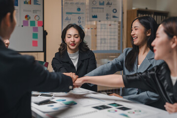 Asian businesspeople joining hands over a table with documents and graphs, celebrating successful teamwork and collaboration in a modern office environment