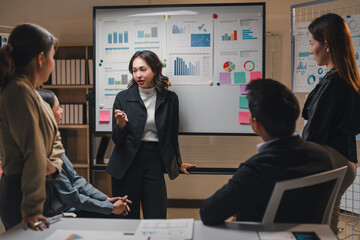 Young asian businesswoman leading a meeting with her colleagues, presenting charts and graphs on a whiteboard in a modern office, demonstrating professionalism and teamwork in a corporate setting