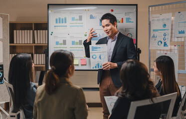 Asian businessman leads a presentation in a modern office, using charts and graphs on a whiteboard and handouts to explain business results to his team of colleagues