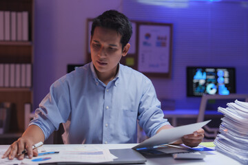 Young businessman reviewing financial reports while working late in a modern office, with a...