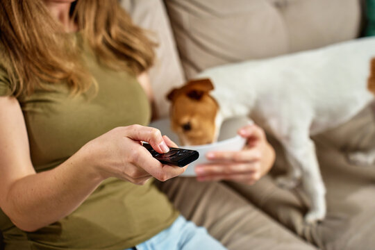 Woman relaxing on sofa with remote control while dog steals food. Pet dog eat food from bowl while owner focuses on TV. Home life and pet behavior concept - Powered by Adobe