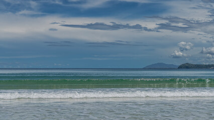 Beautiful seascape. The waves of the turquoise ocean roll towards the shore, foam and spread across the beach. Mountains on the horizon against a background of blue sky and clouds. Malaysia. Borneo.