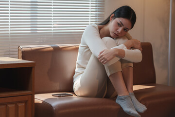 Young asian woman is feeling down and hugging her knees while sitting on the sofa at home,...