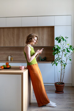Escaping reality woman with poor posture slouched with phone in hand stands in kitchen. Lost in scrolling, screen time habits. Social media addiction of female procrastinating with smartphone at home 