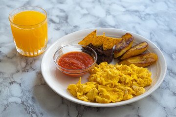 Breakfast of fried eggs, beans with nachos, cream, fried plantains, chirmol, orange juice, on a marble surface
