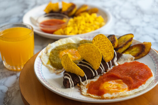 Photograph of two breakfasts with fried eggs, beans with nachos, cream, fried plantains, chirmol, orange juice, on a wooden base and marble surface