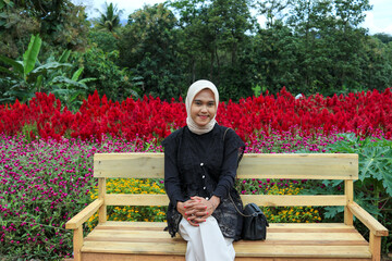 A young woman wearing a hijab and casual outfit sits on a simple wooden park bench.