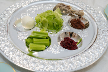 A silver Seder plate is artfully arranged with symbolic foods for Passover. The plate features a roasted bone, lettuce, parsley, charoset, horseradish, and an egg, integral to the celebration.