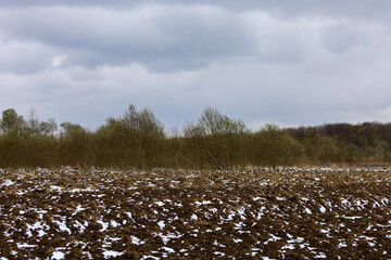 A snow-covered field after plowing stretches to a treeline on the horizon under an overcast, gray...