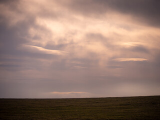 Open field at dusk with dramatic clouds subtly resembling a human face. Warm sunset light adds a mystical, dreamlike atmosphere to the sky.