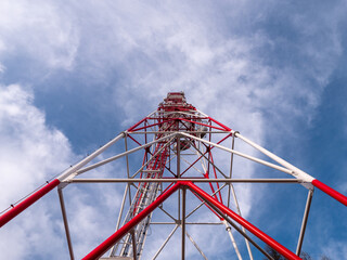 Low-angle view of a red and white telecom tower with antennas and satellite dishes, rising into a partly cloudy blue sky. Modern communication structure meets dynamic sky.