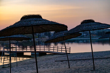 picturesque sunset over a calm lake. reed beach umbrellas standing on a sandy beach. In the background you can see a jetty and the pastel colors of the sky reflected in the water. The whole thing crea