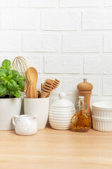 Kitchen utensils, basil plant and condiments resting on wooden countertop in front of white brick wall