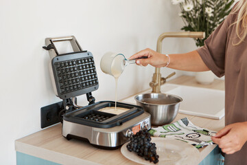 In a beautiful stylish kitchen, a woman bakes waffles close-up. There are fresh flowers, a golden faucet and fruits on the background. Healthy breakfast, family happiness, mother's love. High quality