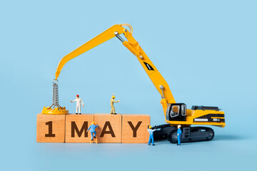 The word 1 MAY is written on a wooden box, miniature people and excavator trucks make up the box.