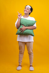 Calm Asian Man in Pajamas and Sleep Eye Mask Hold Pillow and Point Index Finger Aside to Copy Space