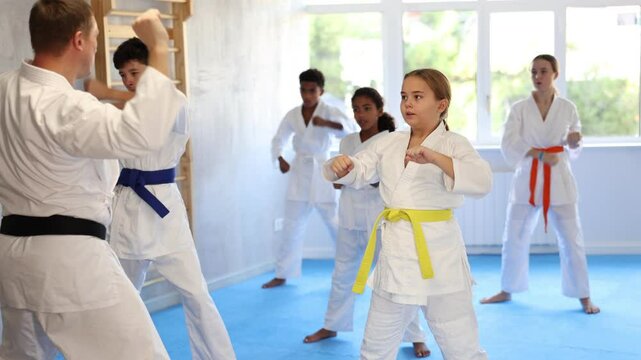 Concentrated girl in white kimono practicing punches in gym during martial arts workout with multiracial group of tweens. Shadow fight, combat sports training concept 