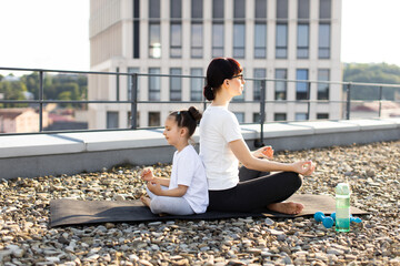 Caucasian adult woman and young girl sitting back-to-back meditating outdoors together on a rooftop. They practice yoga and mindfulness during a sunny day.