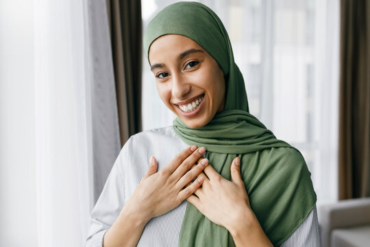 Indoor portrait of grateful beautiful young muslim woman in stylish green headscarf pressing her palms against chest expressing gratitude and respect, looking at camera with adorable smile