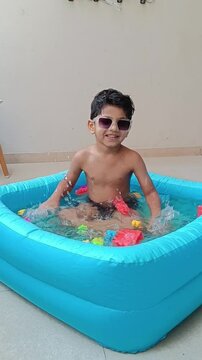 4 year old cute, young Indian boy relaxing in a kiddie pool- bath tub and splashing water, wearing stylish sunglasses and enjoying a fun indoor summer activity during summer vacation.