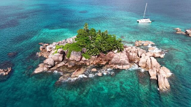 Praslin Island, Seychelles, featuring a tiny rocky islet with palm trees surrounded by crystal-clear turquoise ocean and a nearby sailboat.

