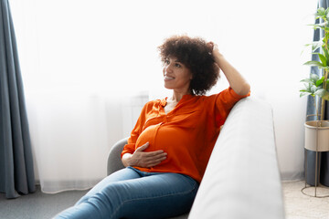 Fototapeta premium Smiling pregnant African American woman relaxing on sofa and touching belly
