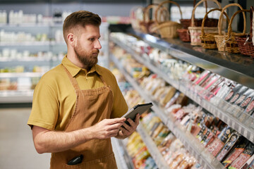 Bearded store employee wearing apron checking inventory on digital device while standing in front...