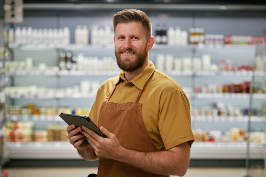 Portrait of friendly grocery store employee holding tablet and smiling while standing in front of store shelves filled with various products