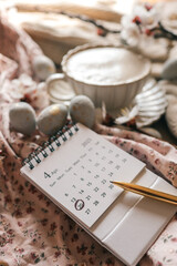 Close-up of an April 2025 calendar with April 20 marked, lying on a floral fabric beside speckled Easter eggs, a cup of coffee, and a golden pen in warm natural light