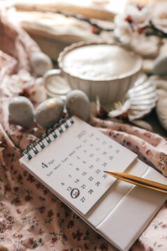 Close-up of an April 2025 calendar with April 20 marked, lying on a floral fabric beside speckled Easter eggs, a cup of coffee, and a golden pen in warm natural light