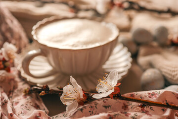 Cup of frothy coffee in an elegant vintage-style saucer, surrounded by cherry blossoms, pastel Easter eggs, and soft fabrics — cozy springtime still life composition