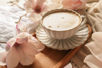 Elegant cup of cappuccino on a white saucer with blooming magnolia flowers on a wooden tray in soft morning light