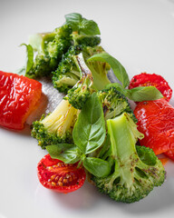 A close-up of a healthy vegetable dish featuring roasted broccoli, cherry tomatoes, and red bell peppers, garnished with fresh basil leaves. 