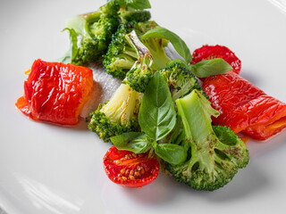A close-up of a healthy vegetable dish featuring roasted broccoli, cherry tomatoes, and red bell peppers, garnished with fresh basil leaves. 