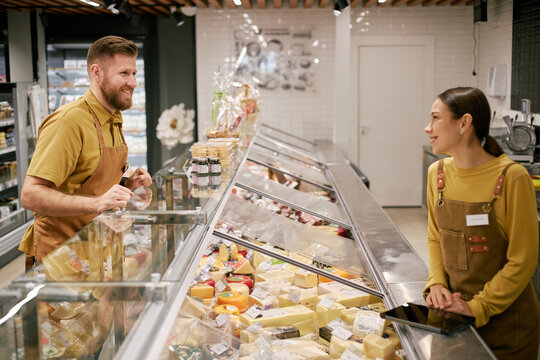 Male shop assistant with beard and female customer discussing cheese options while standing at deli counter. Both smiling and engaging in friendly conversation, display of assorted cheeses visible