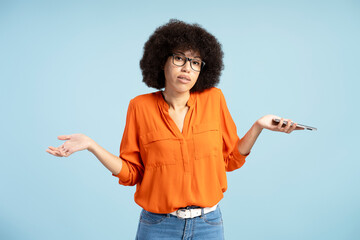 Puzzled young woman shrugging shoulders holding smartphone posing against blue background