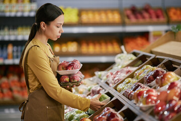 Woman arranging fresh fruits in grocery store display holding apples inspecting pears ensuring...