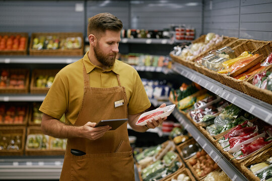 Male grocery store worker checking packaged meat holding digital tablet. Store shelves filled with various fresh products in background indicating well-stocked store